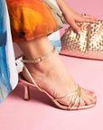 Gold high-heeled sandal worn on a pink surface with a colorful dress and gold clutch in the background