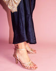 Gold high-heeled sandals worn with navy dress on a pink background