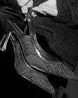 Black high-heeled shoes with intricate design held by a person against a dark background