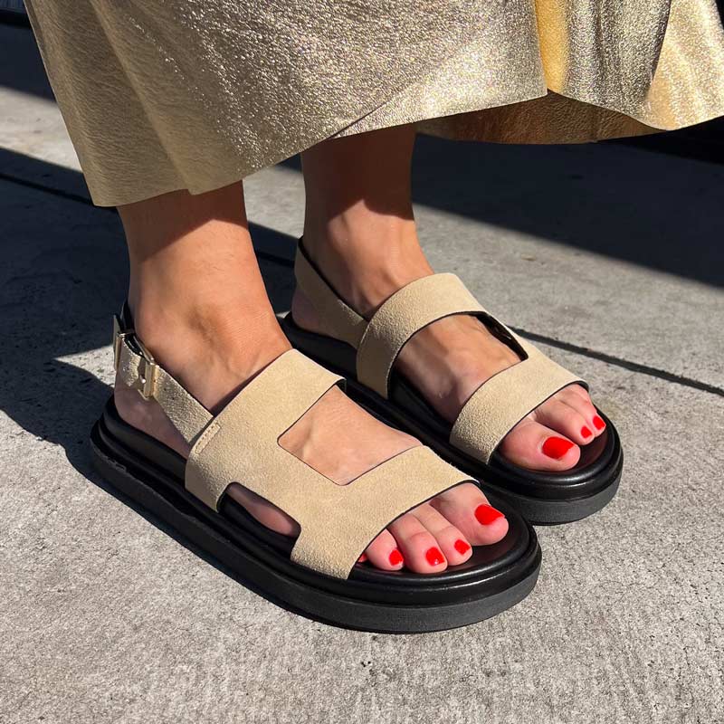 Beige sandals with black soles worn by a person on a concrete surface.