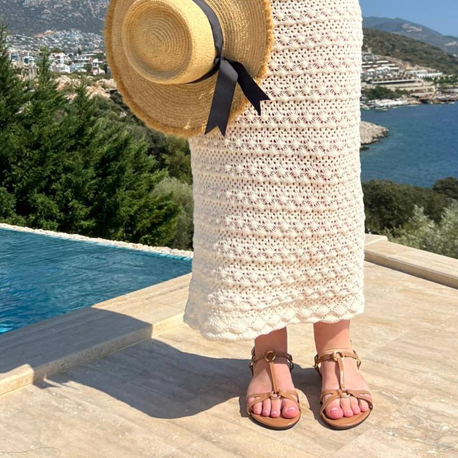 Person wearing a straw hat, white dress, and sandals by a pool with a scenic background.