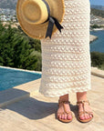 Person wearing a straw hat, white dress, and sandals by a pool with a scenic background.