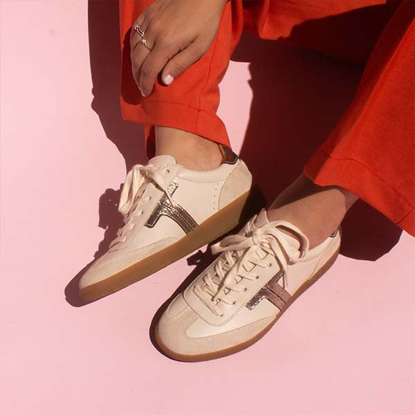 model sitting on ground wearing bright orange pants and cream leather sneaker sneakers with gum sole in front of pink background