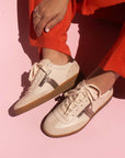 model sitting on ground wearing bright orange pants and cream leather sneaker sneakers with gum sole in front of pink background