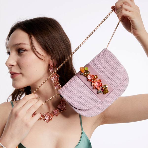 Model wearing green dress and statement flower necklace and earrings holding NALÌ CARLA clutch in lilac with 3D flower embellishments and gold shoulder chain on white background