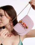 Model wearing green dress and statement flower necklace and earrings holding NALÌ CARLA clutch in lilac with 3D flower embellishments and gold shoulder chain on white background