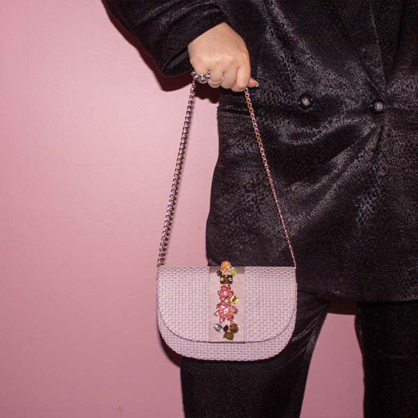 model wearing balck satin suit in front of pink wall holding NALÌ CARLA clutch in lilac with 3D flower embellishments and gold shoulder chain