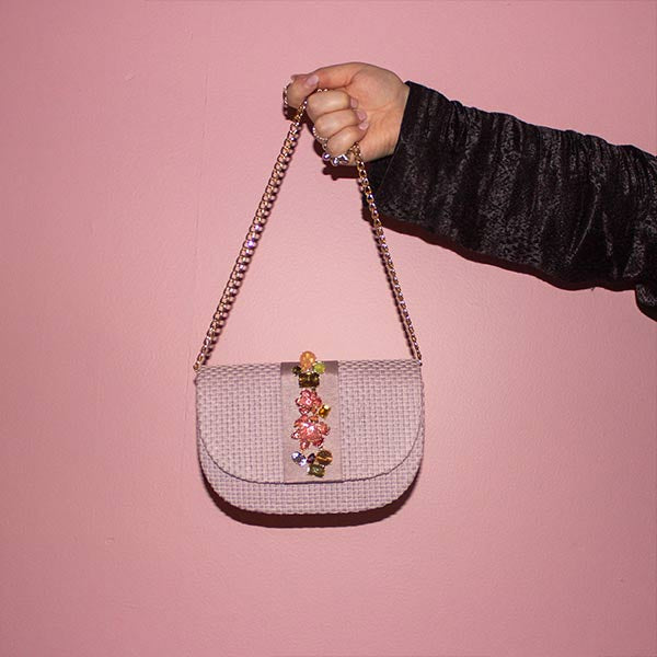 Arm stretched out in front of pink wall holding NALÌ CARLA clutch in lilac with 3D flower embellishments and gold shoulder chain