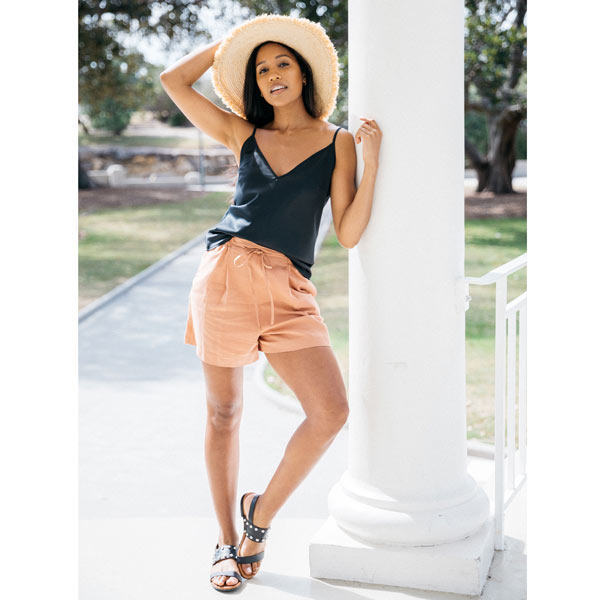 Black leather Billi Bi flat sandal with silver stud detail on model in shorts, cami, sunhat leaning against white column