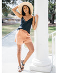 Black leather Billi Bi flat sandal with silver stud detail on model in shorts, cami, sunhat leaning against white column