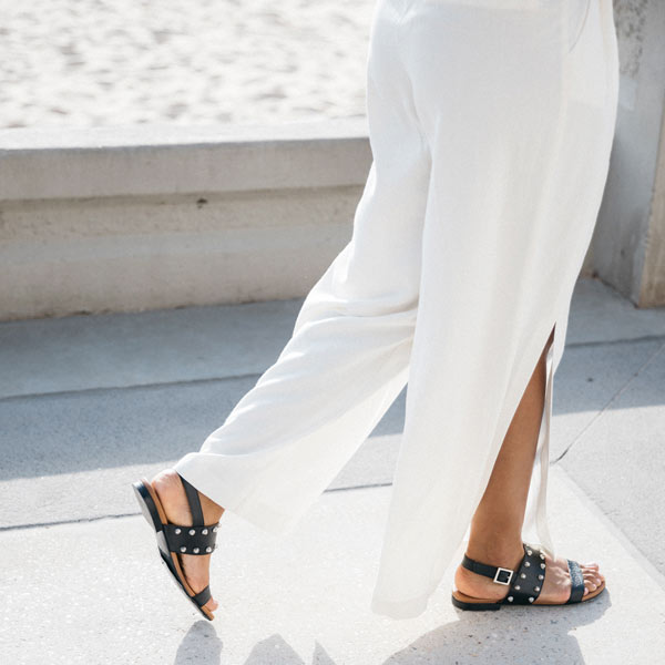 Black textured leather Billi Bi flat sandal with silver stud detail on model walking in white linen pants