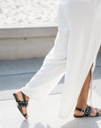 Black textured leather Billi Bi flat sandal with silver stud detail on model walking in white linen pants